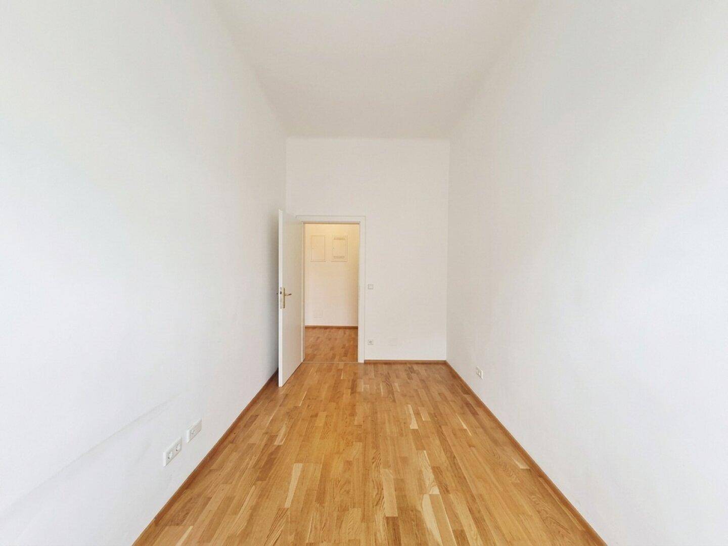 Einfacher, heller Raum mit Parkettboden und Zugang zu einem weiteren Zimmer.