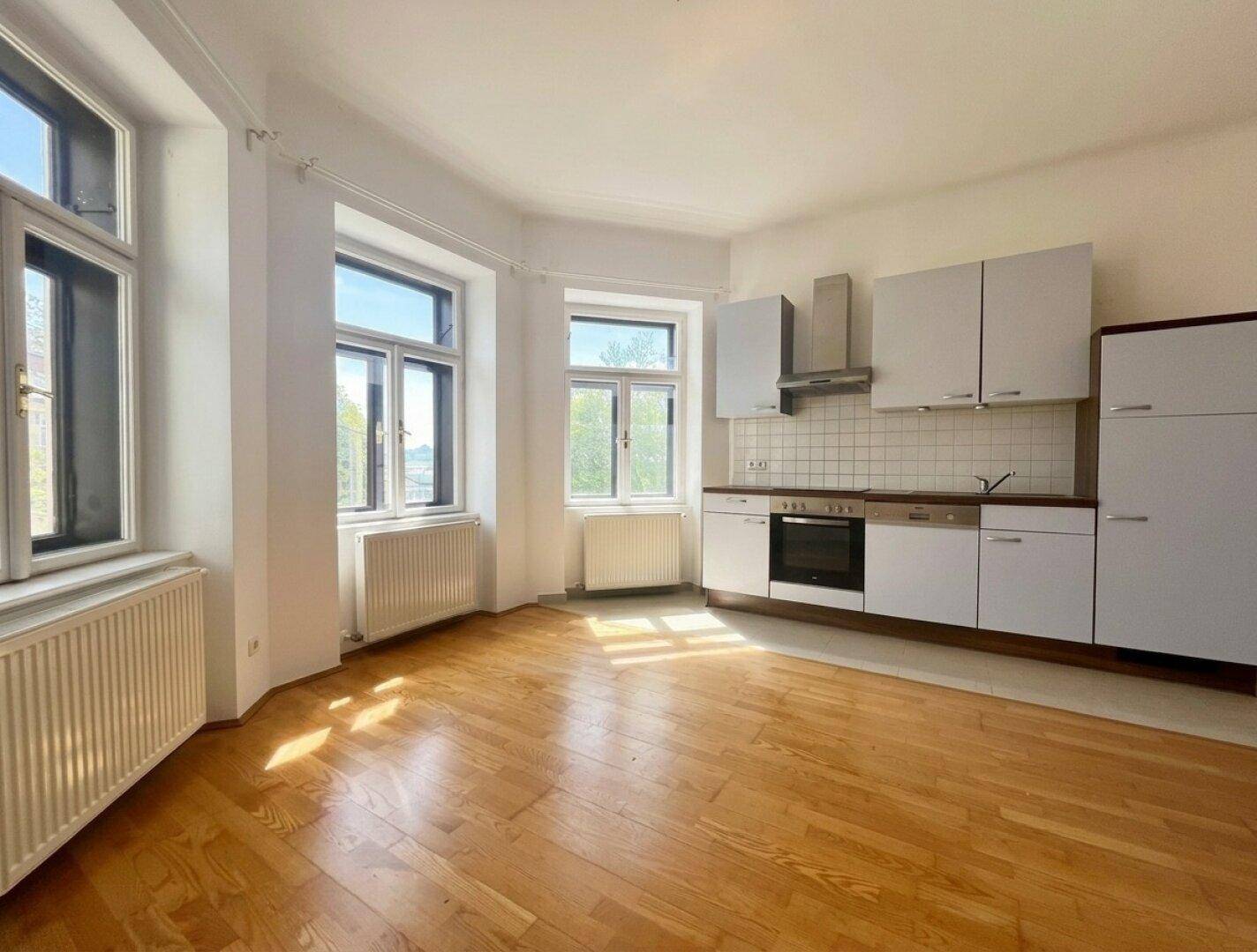 Geräumiger Wohnbereich mit integrierter Küche, Holzboden und viel natürlichem Licht durch die Fenster.