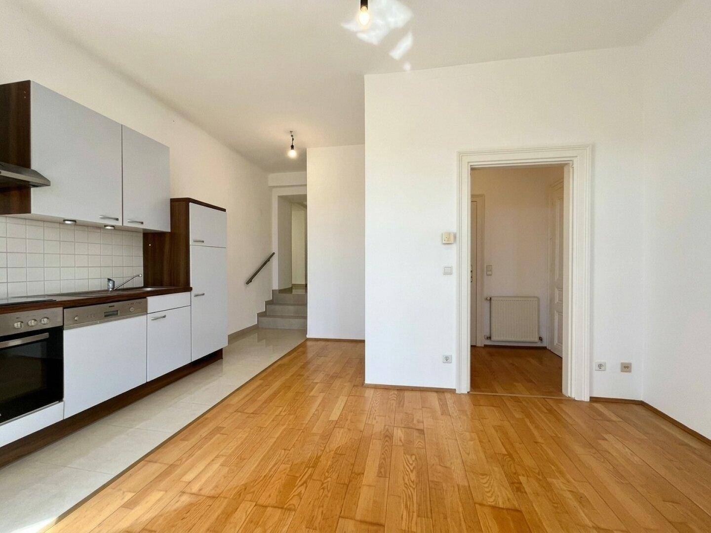 Offene Küche mit Holzboden, weißen Schränken und Zugang zu einem Flur mit Treppe.