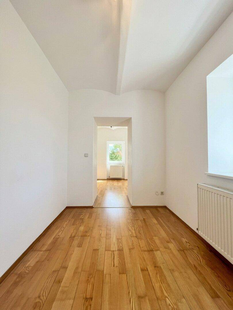 Geräumiges Zimmer mit Holzboden, weißen Wänden und einem Durchgang zu einem weiteren Raum.