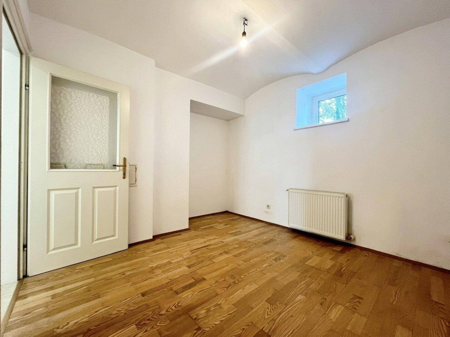 Gemütliches Zimmer mit Holzboden, weißen Wänden und einem kleinen Fenster, ideal als Schlafzimmer.