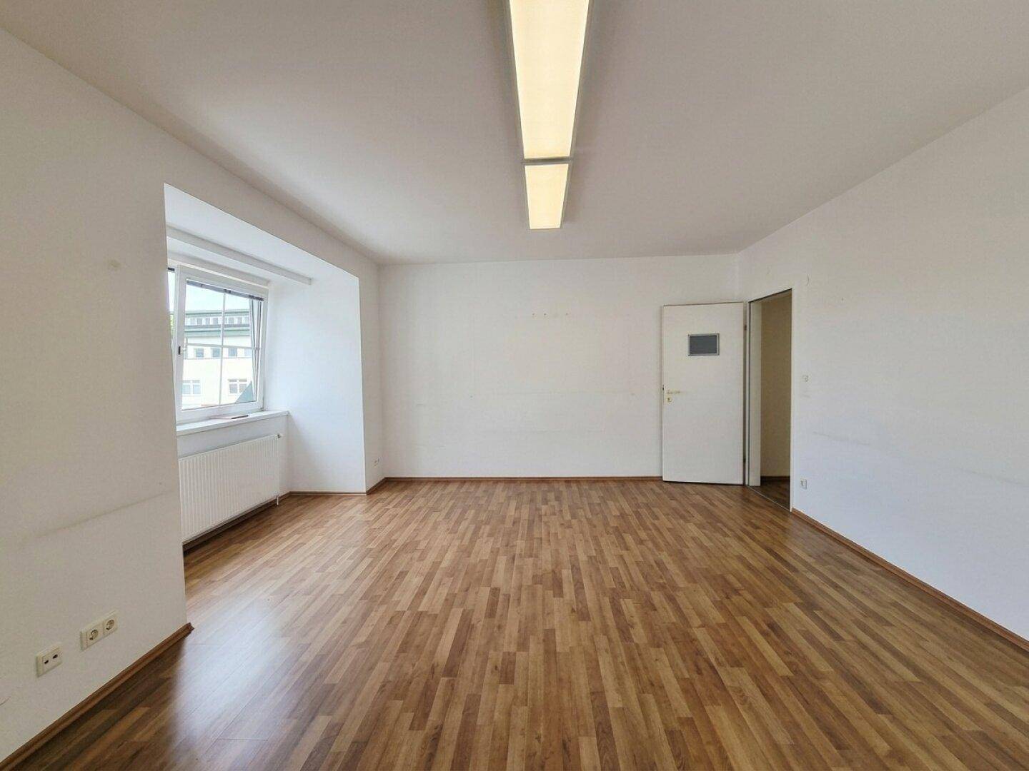 Leeres Zimmer mit Laminatboden, weiß gestrichenen Wänden und einer Tür, die zu einem weiteren Raum führt.