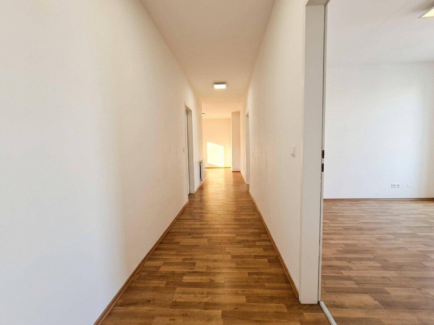 Langer Flur mit Laminatboden und weißen Wänden, der zu mehreren Zimmern führt und hell beleuchtet ist.