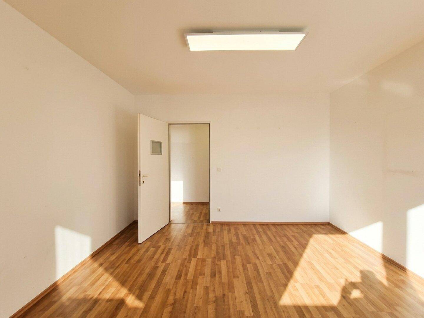 Leeres Zimmer mit Laminatboden, weißen Wänden und einer offenen Tür, die viel Licht hereinlässt.