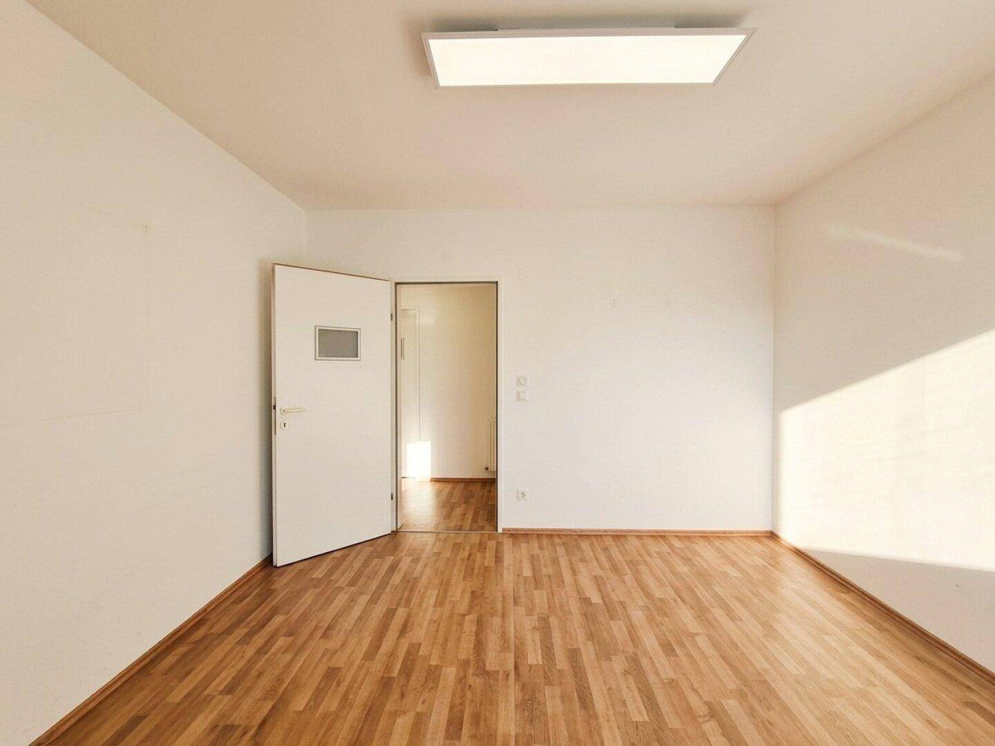 Leeres Zimmer mit Laminatboden, weißen Wänden und einer offenen Tür, die den Blick in den nächsten Raum freigibt.