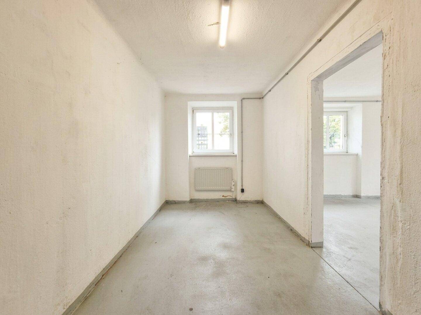 Schmaler Lagerraum mit Betonboden, Fenster und Zugang zu einem angrenzenden Bereich im Keller.