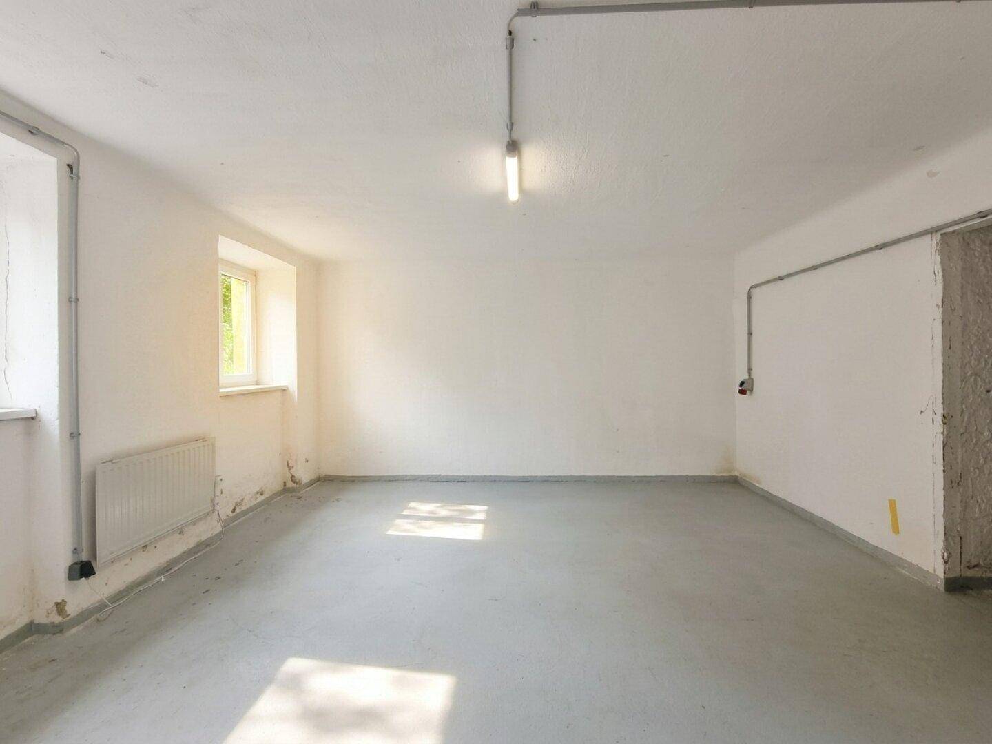 Einfacher Lagerraum mit Betonboden, Heizkörper unter dem Fenster und sichtbaren Leitungen an der Decke.