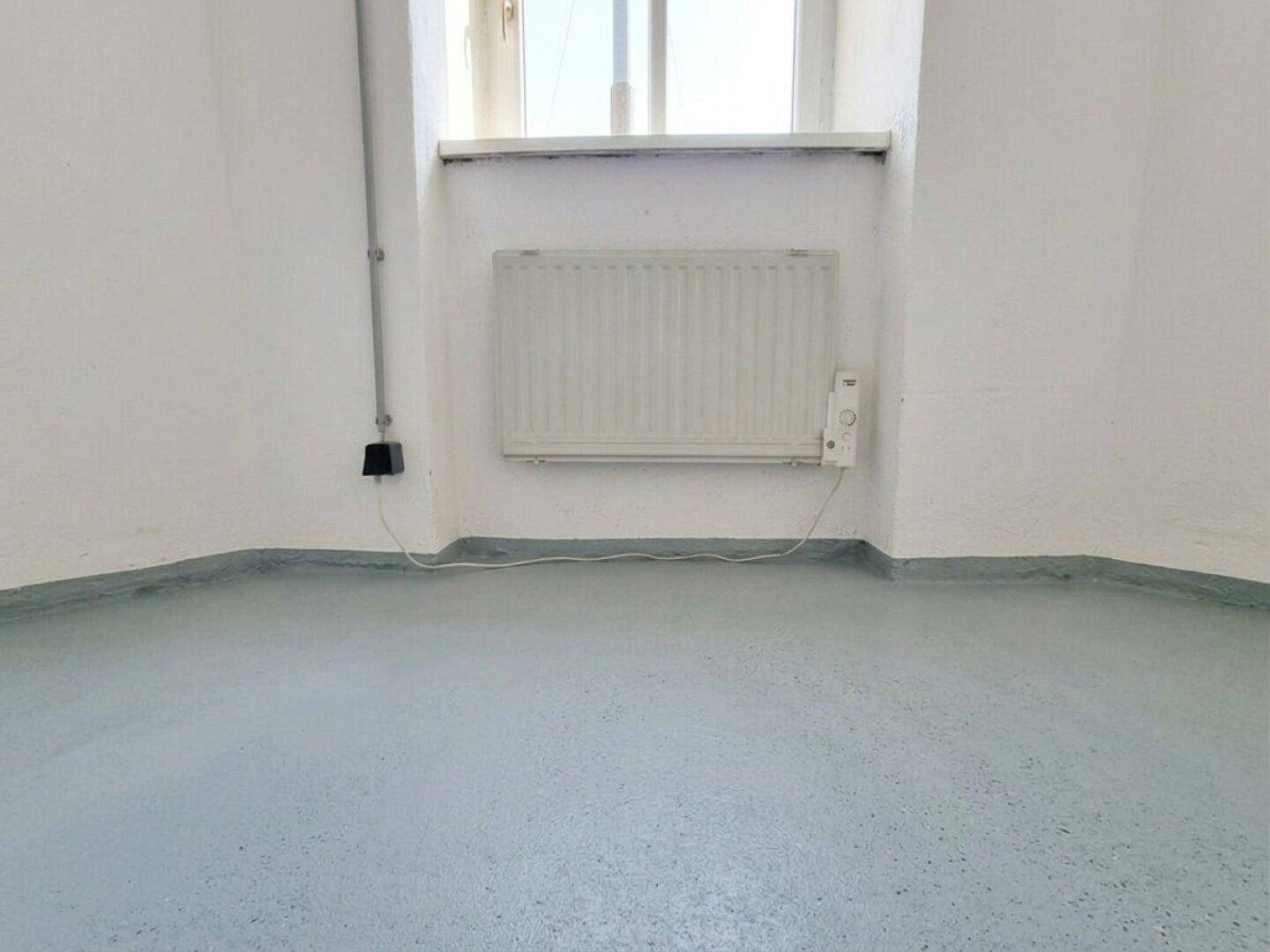 Detailansicht eines Fensters mit Heizkörper und hellblauem Bodenbelag in einem Kellerraum.