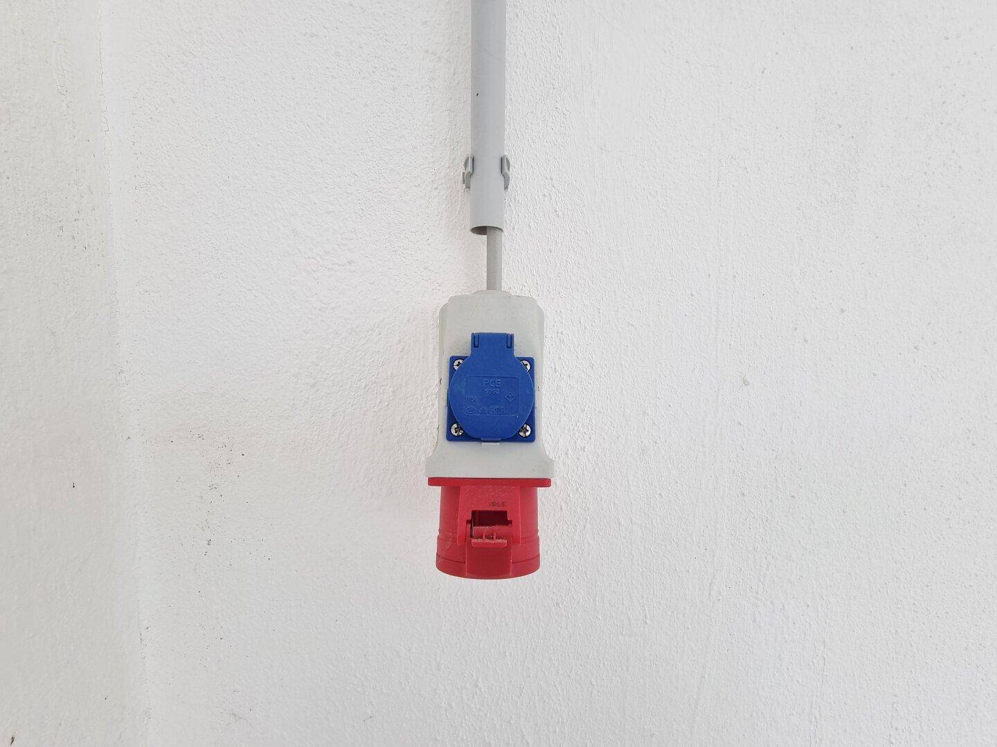 Nahaufnahme einer roten und blauen Industriesteckdose an einer weißen Wand mit sichtbaren Leitungen.