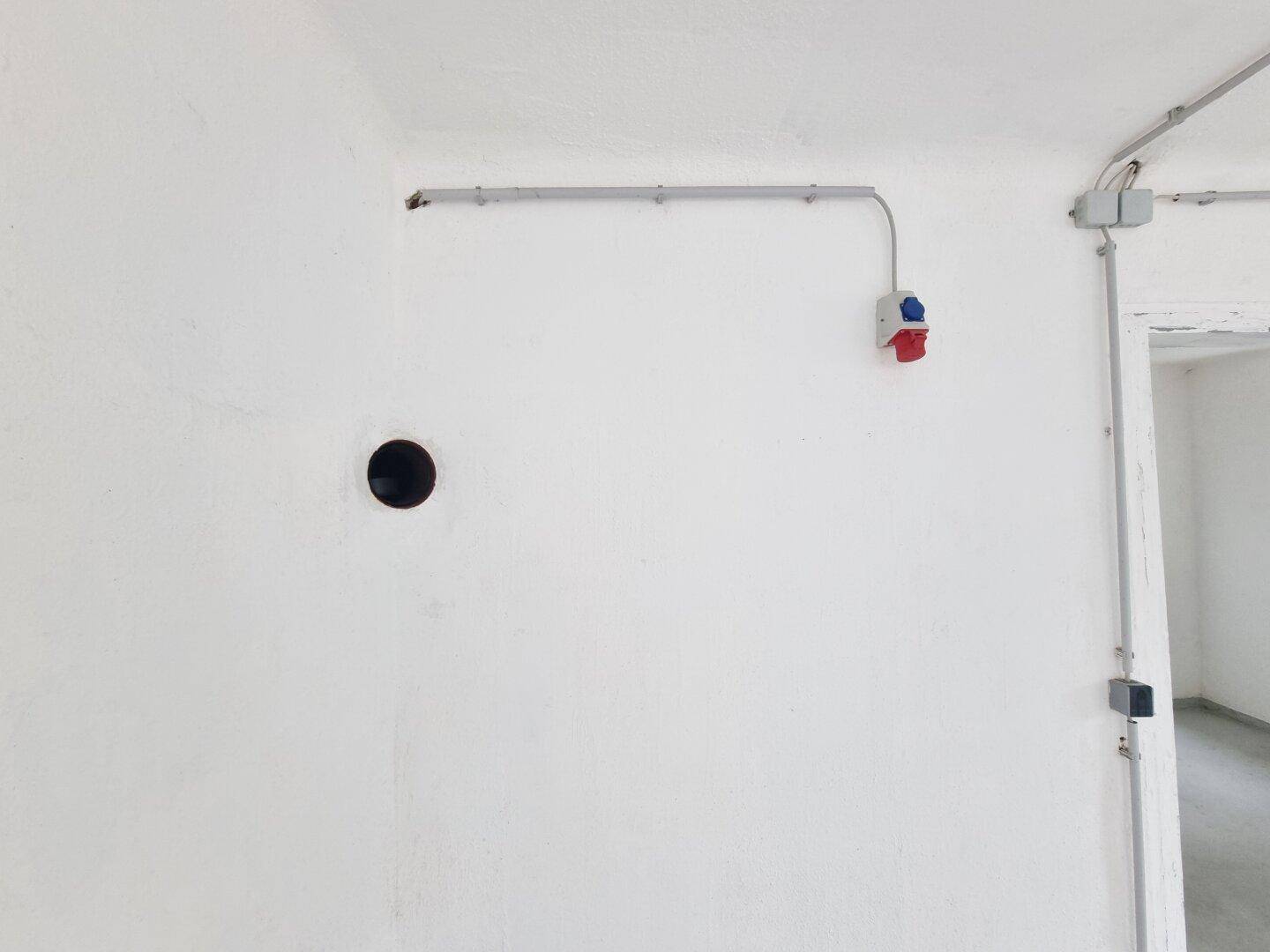Weiße Wand mit sichtbaren Leitungen und roten sowie blauen Industriesteckdosen.