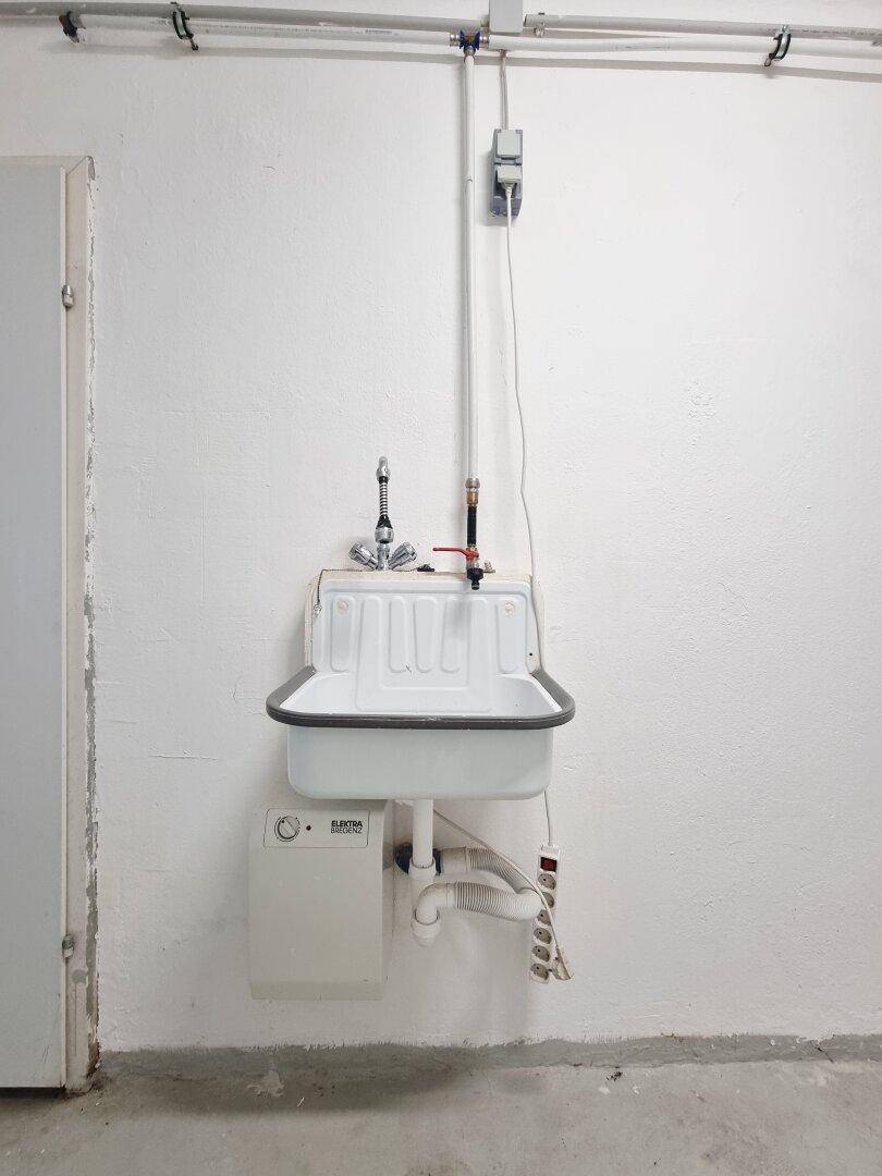 Nahaufnahme eines Waschbeckens mit Warmwasserbereiter und sichtbaren Installationen an der Wand.