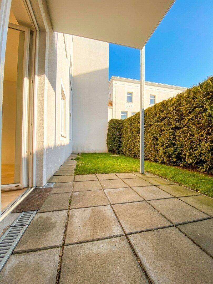Sonniger Terrassenbereich mit Steinplatten und Blick auf die grüne Hecke und das Nachbargebäude.