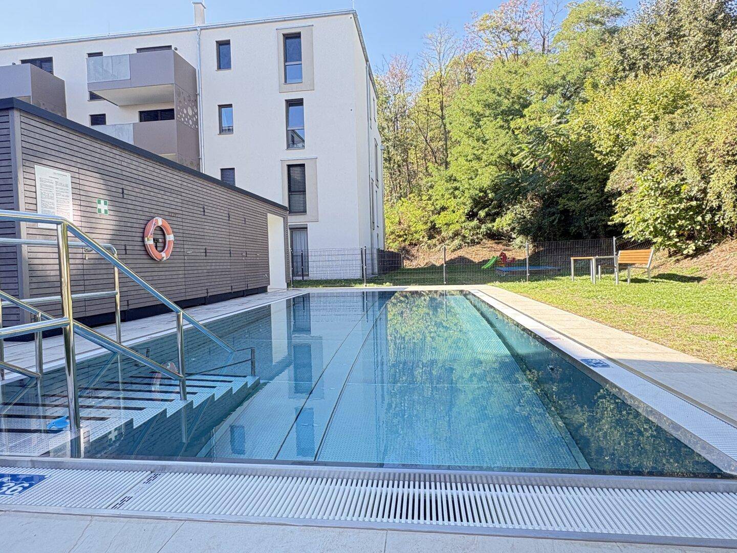 Außenpoolbereich mit modernem Gebäude im Hintergrund und umgebender Grünfläche.