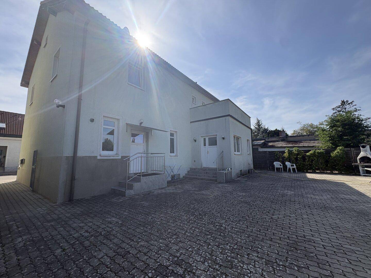 Außenansicht des Gebäudes mit gepflastertem Vorplatz und Sonnenschein, ideal für Grillabende.
