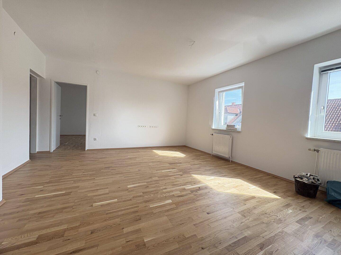 Geräumiges Wohnzimmer mit hellem Holzparkettboden und großen Fenstern, lichtdurchflutet.