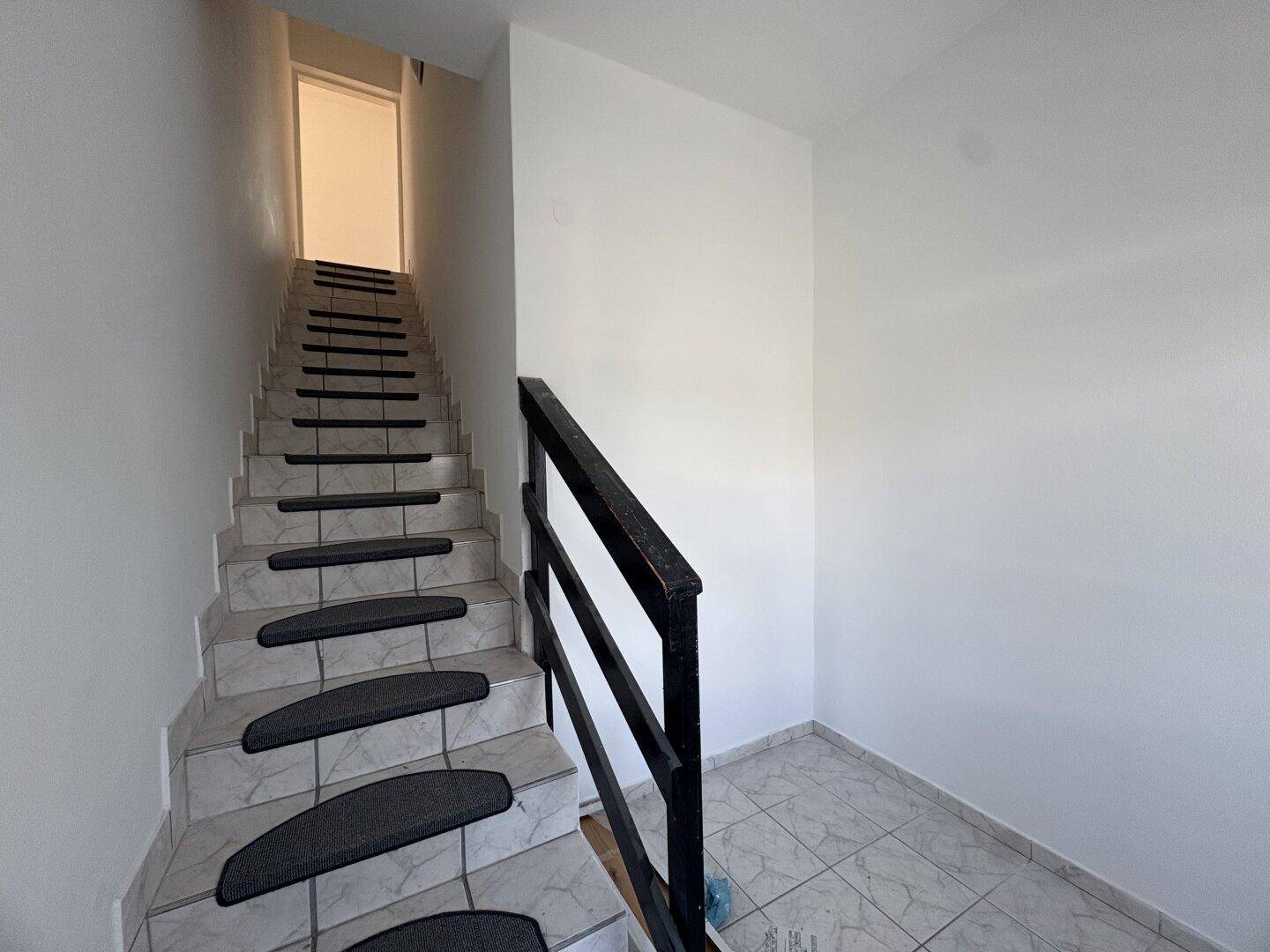 Treppenhaus mit gefliesten Stufen und schwarzem Geländer, das in die obere Etage führt.