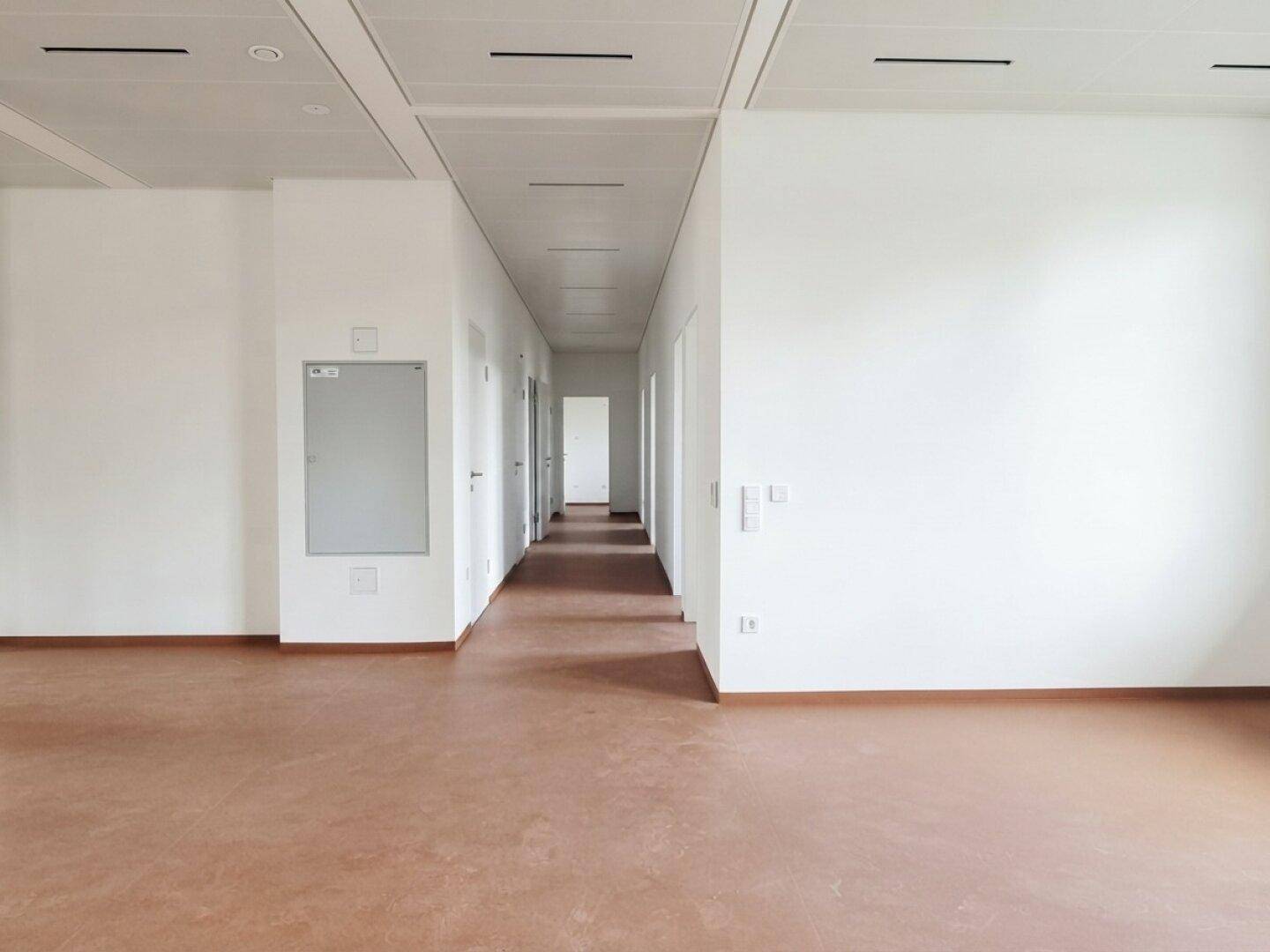 Langer, heller Flur mit braunem Boden und weißen Wänden in einem modernen Gebäude.