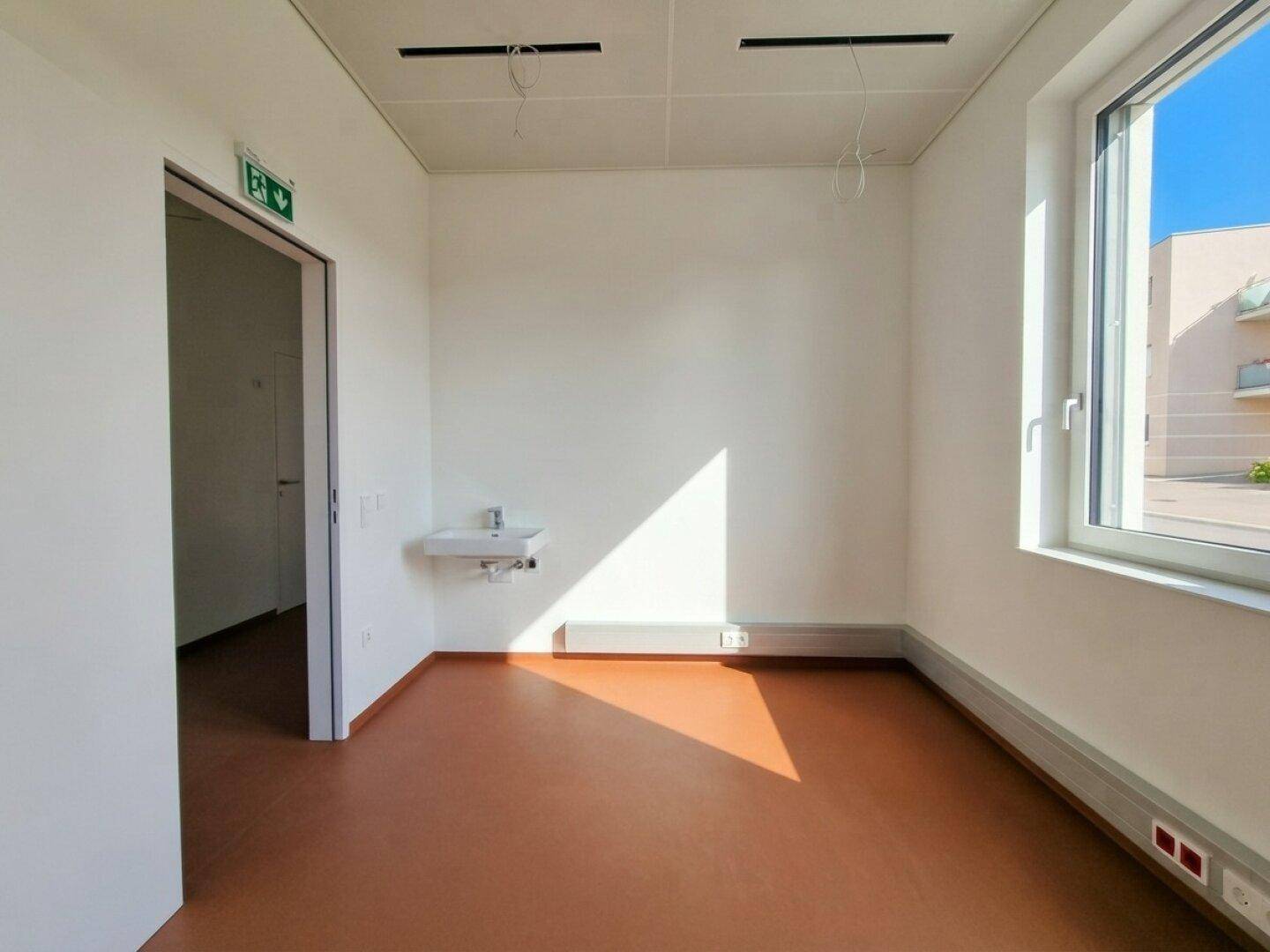 Kleiner, heller Raum mit Fenster, Waschbecken und braunem Boden, geeignet als Behandlungszimmer.