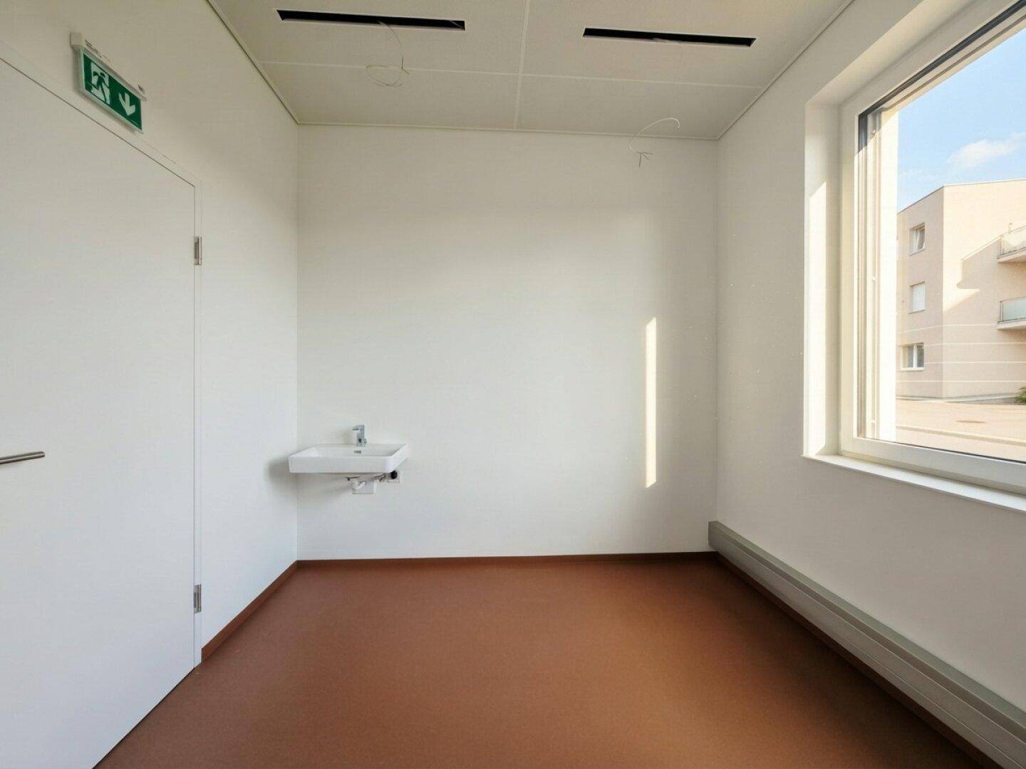 Heller Raum mit Fenster, Waschbecken und braunem Boden, ideal für ein kleines Büro oder Wartezimmer.