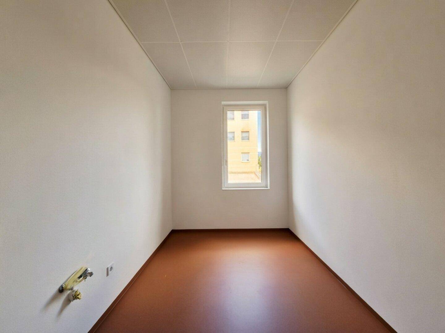 Schmaler, heller Raum mit einem Fenster und braunem Boden, geeignet als kleines Büro oder Abstellraum.