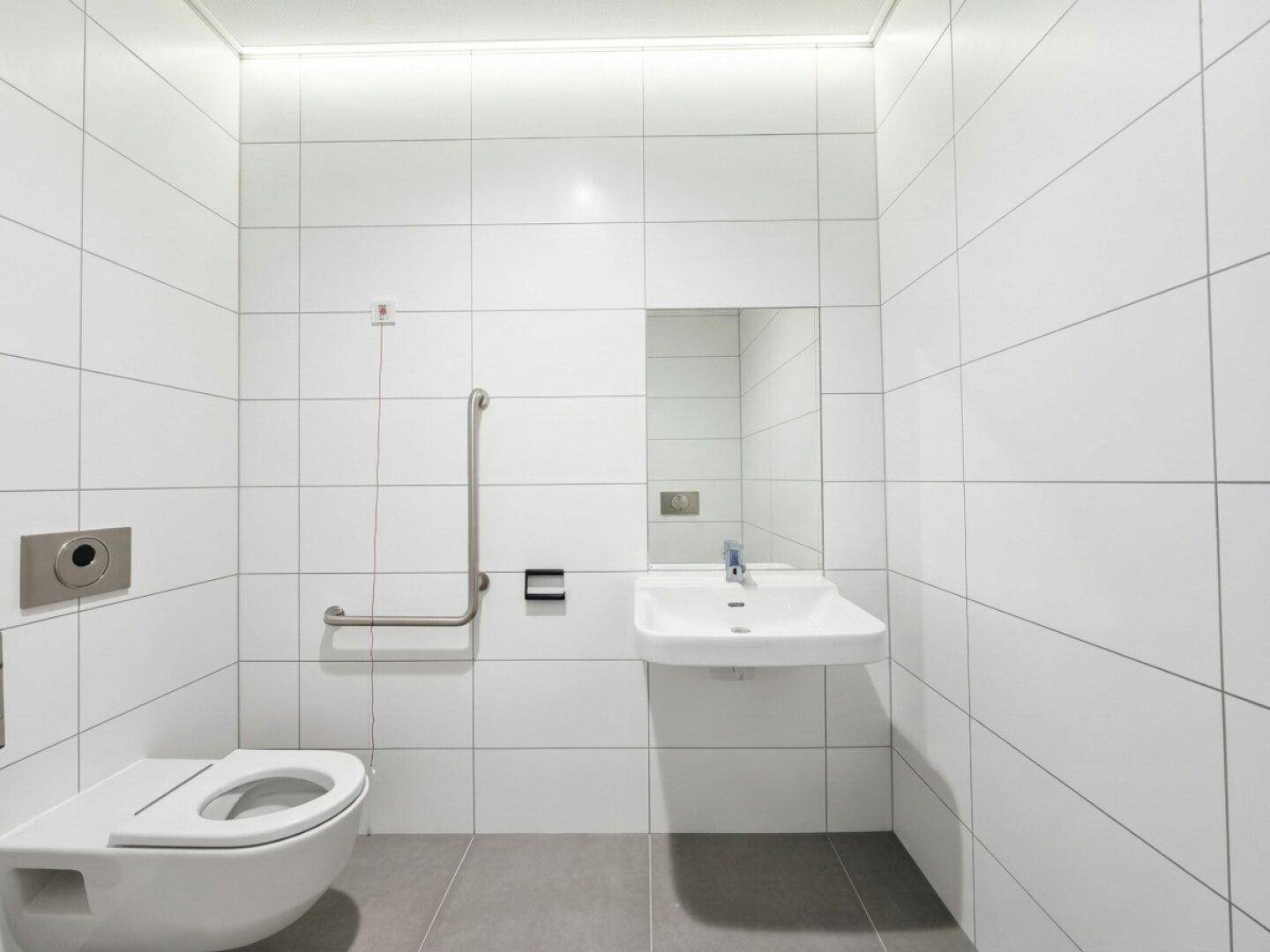 Modernes, barrierefreies Badezimmer mit weißen Fliesen, Toilette, Waschbecken und Haltegriffen.