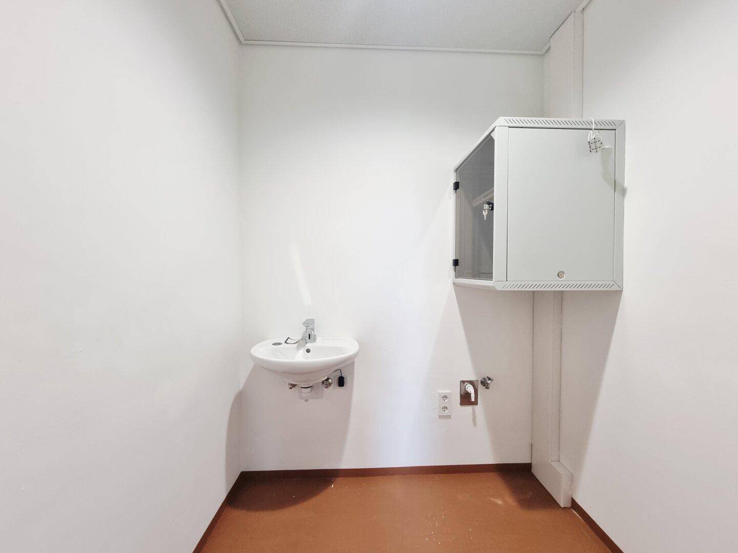 Kleiner Raum mit Waschbecken, Schrank und braunem Boden, möglicherweise ein Abstell- oder Putzraum.