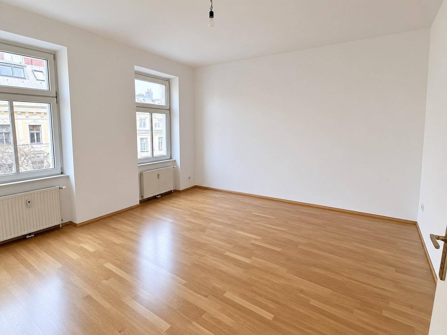 Geräumiges Zimmer mit hellem Parkettboden und zwei Fenstern, die viel Tageslicht hereinlassen.
