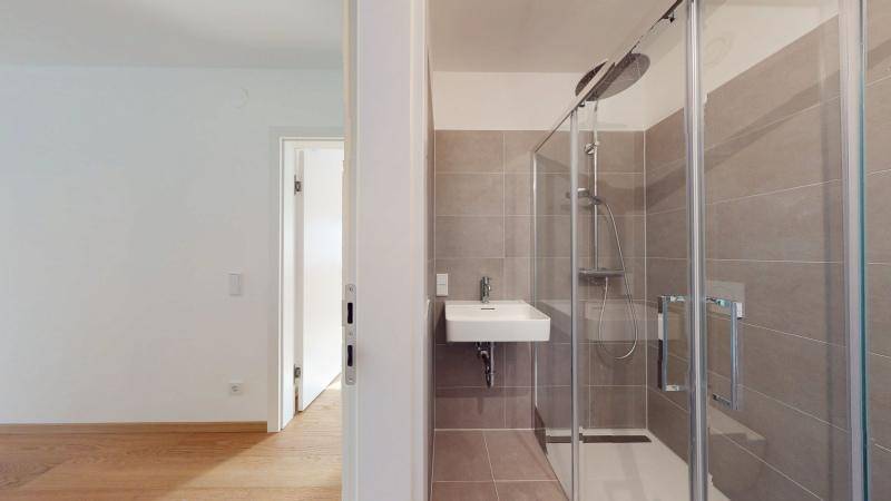 Modernes Badezimmer mit Duschkabine und grauen Fliesen, angrenzend an einen weiteren Raum.