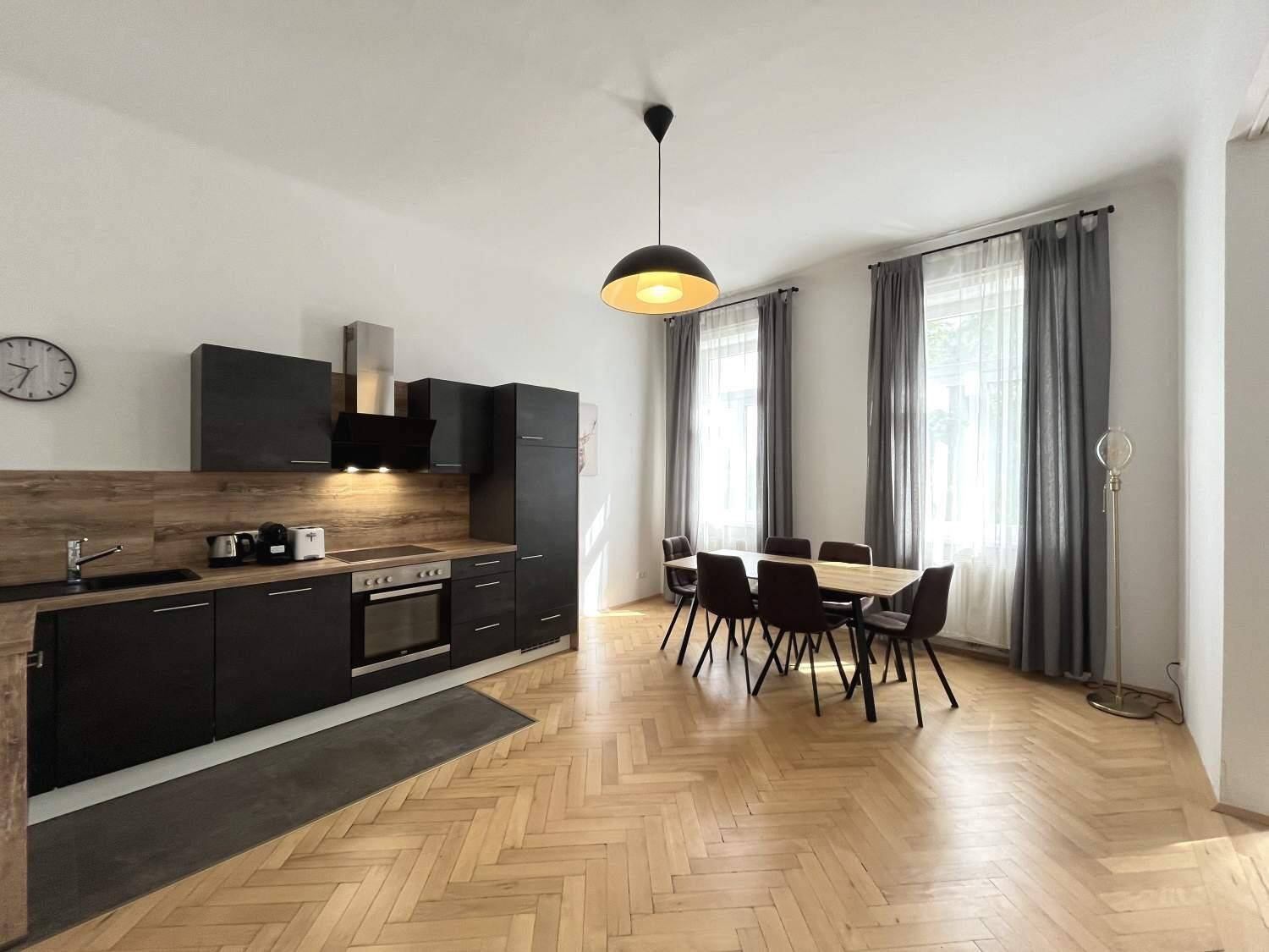 Moderne Küche mit dunklen Schränken und Essbereich, hell durch große Fenster und Parkettboden.