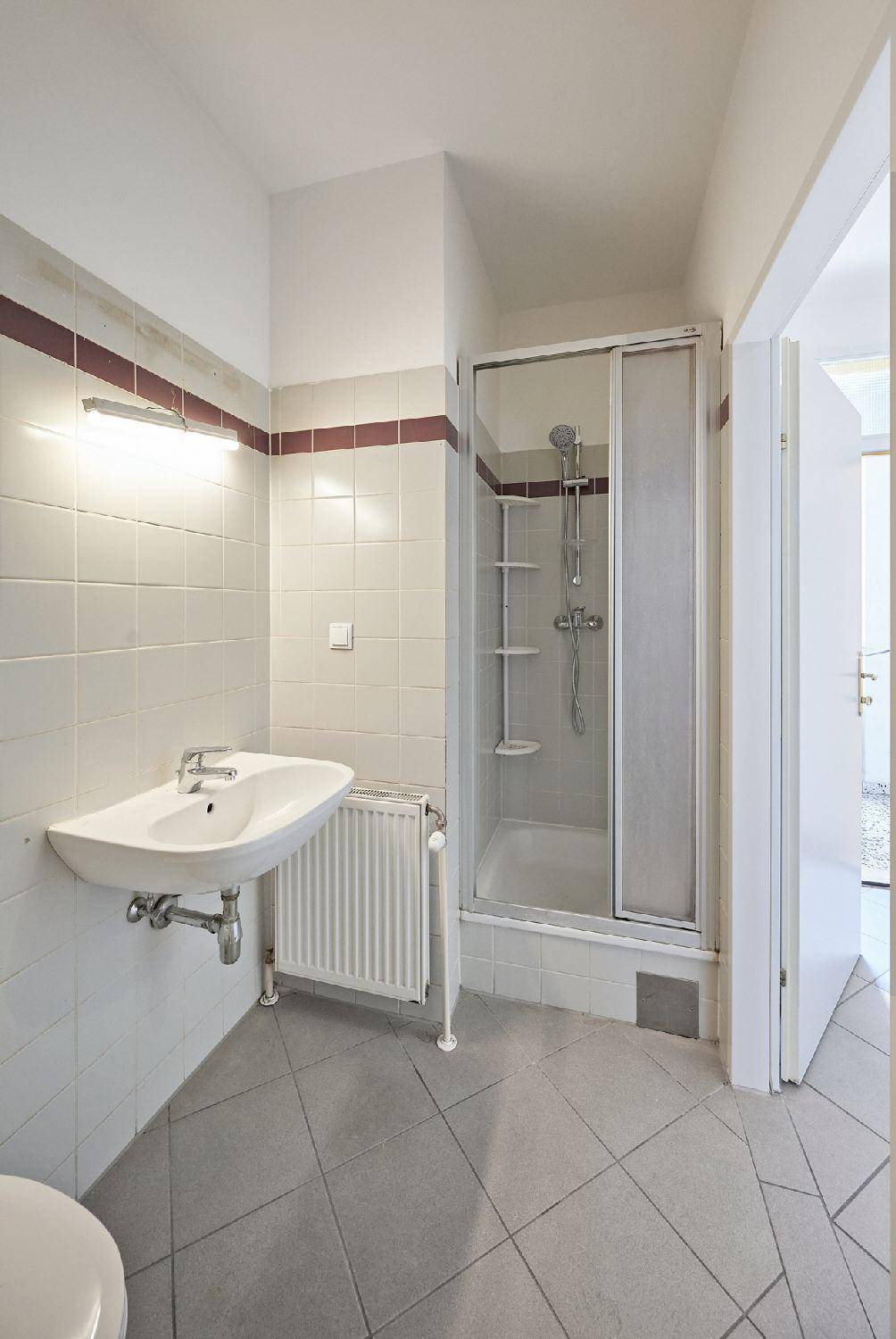 Funktionales Badezimmer mit Duschkabine, Waschbecken und Heizkörper, hell gefliest.