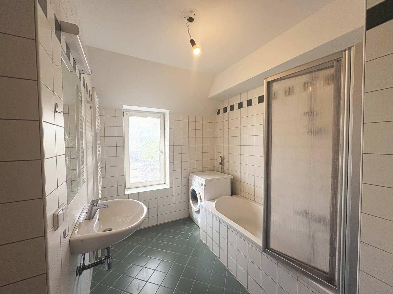 Gefliestes Badezimmer mit Badewanne, Dusche, Waschbecken und Fenster, inklusive Waschmaschine.