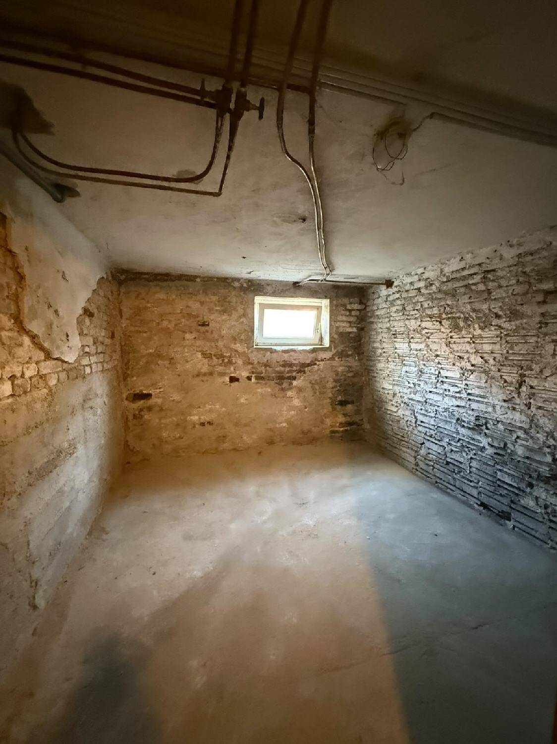 Unfertiger Kellerraum mit unverputzten Ziegelwänden, Betonboden und kleinem Fenster.