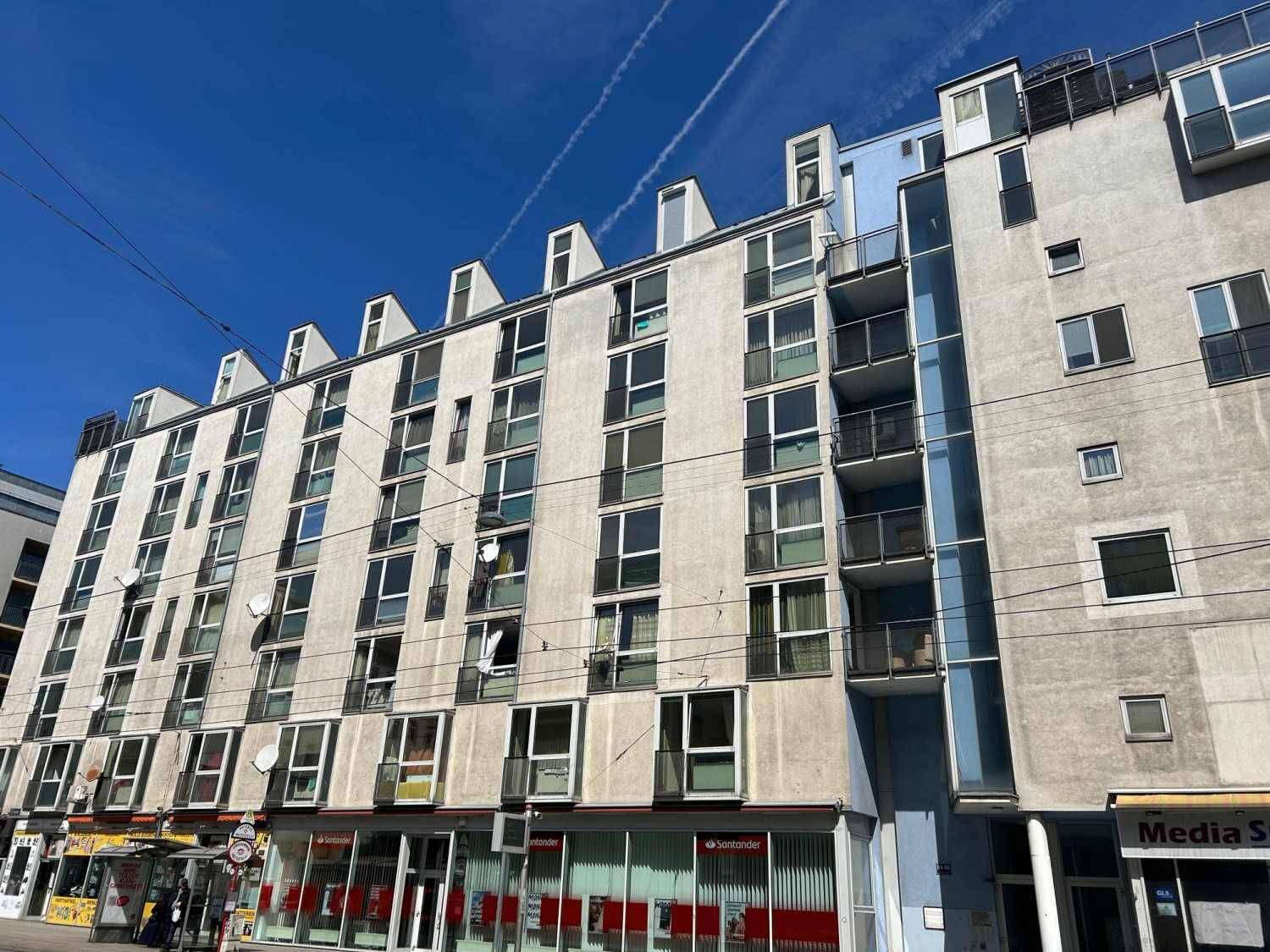 Moderne Wohnanlage mit mehreren Etagen, Balkonen und Geschäftsflächen im Erdgeschoss an einer Straße.