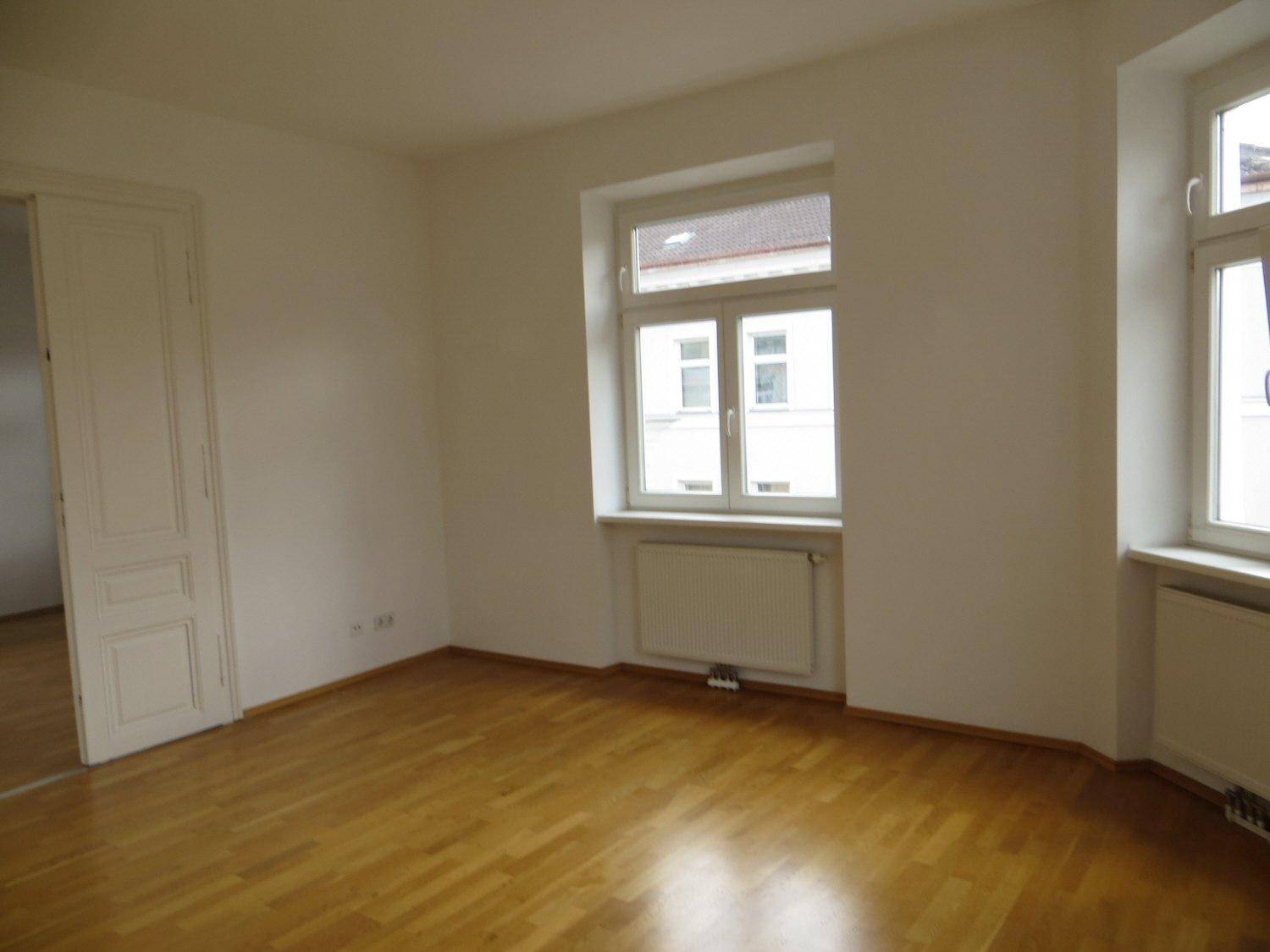 Heller Raum mit Parkettboden, einem großen Fenster und einer weißen Tür.