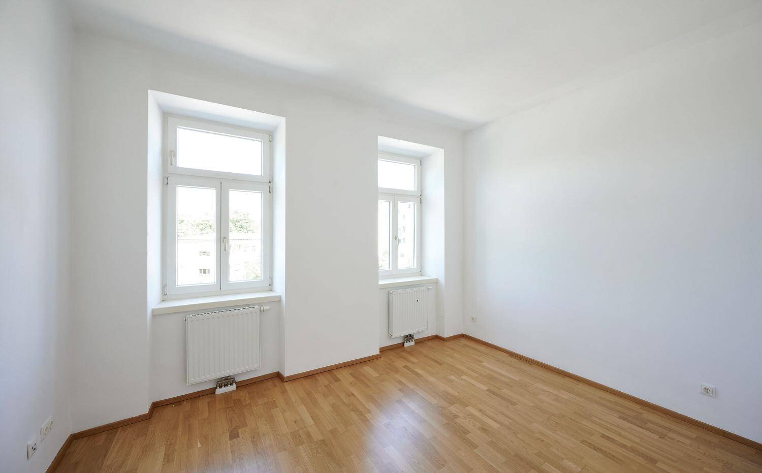 Helles, leeres Zimmer mit Parkettboden und zwei Fenstern, bereit zur individuellen Gestaltung.
