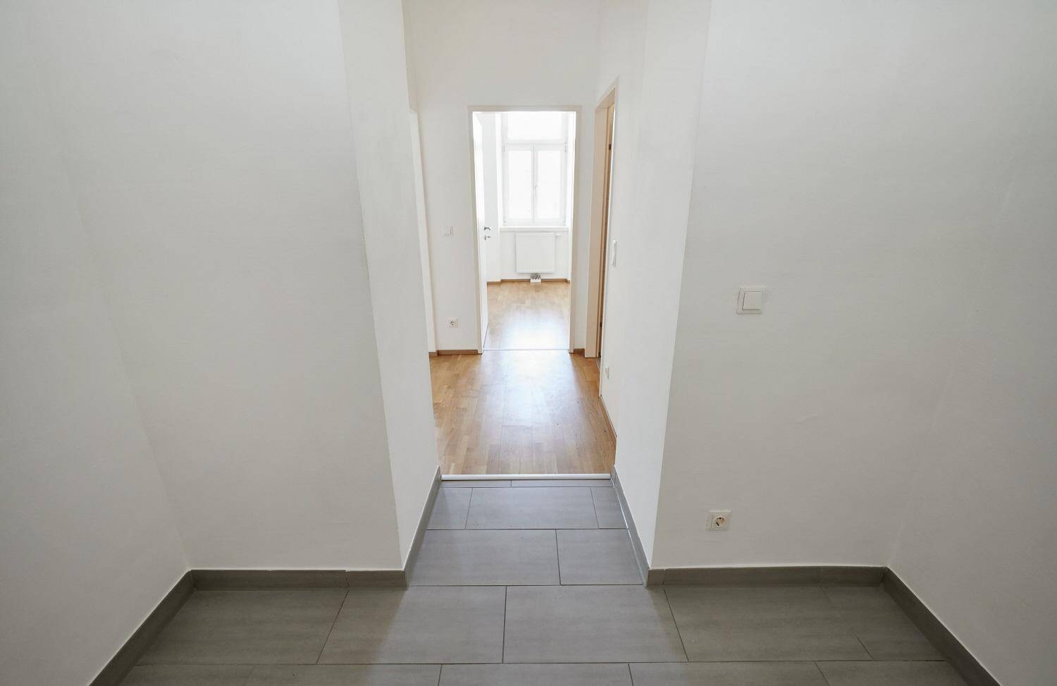 Langer, heller Flur mit Fliesenboden, der zu einem Zimmer mit Parkett führt.