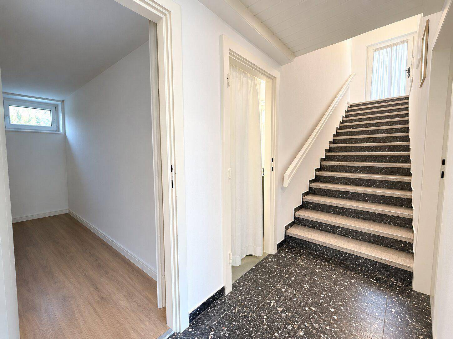 Heller Flur mit Treppenaufgang und dunklem Terrazzoboden, der zu weiteren Räumen führt.
