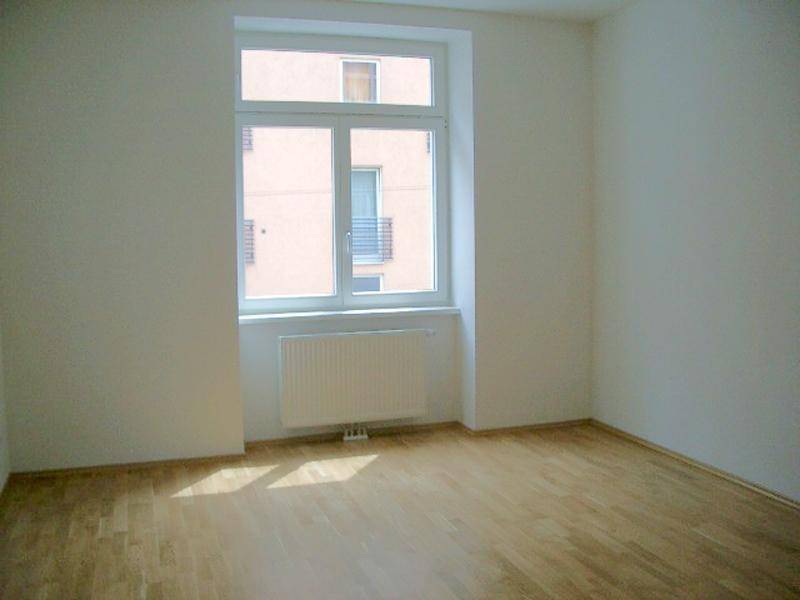 Leeres Zimmer mit Holzboden, großem Fenster und Heizkörper, bereit zur individuellen Gestaltung.