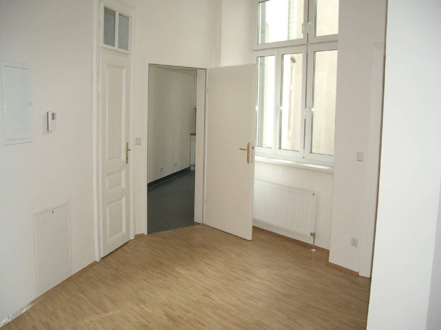 Heller Raum mit Parkettboden und Fenster, eine offene Tür führt zu einem angrenzenden Bereich.