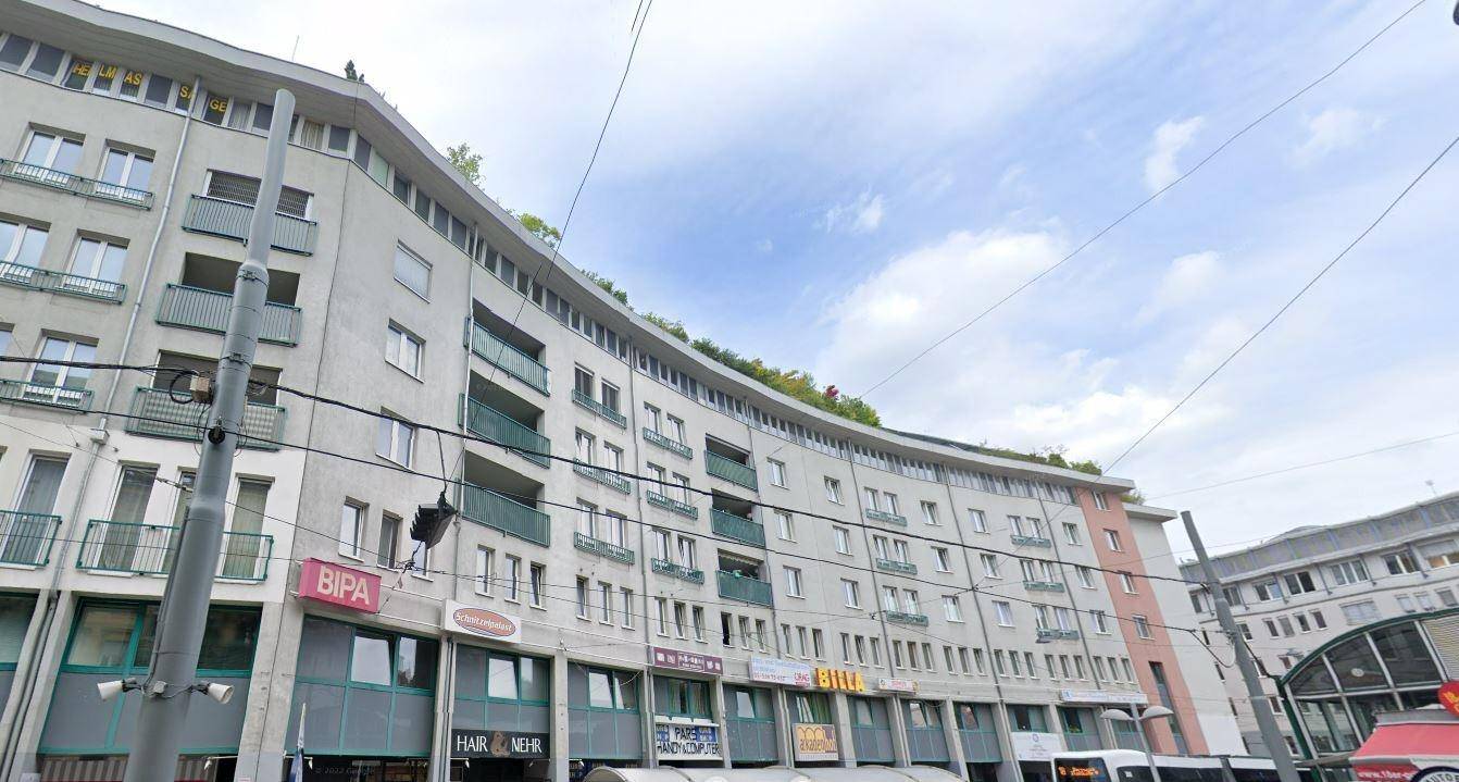 Mehrstöckiges Gebäude mit vielen Fenstern, Balkonen und Geschäften im Erdgeschoss.