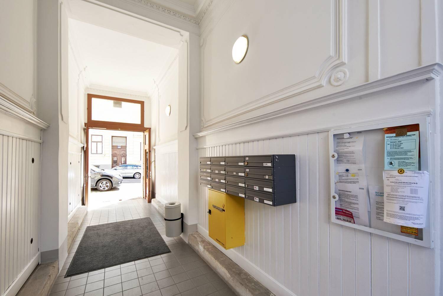 Ein heller Eingangsbereich mit Briefkästen, einer Informationstafel und Blick zur Straße.