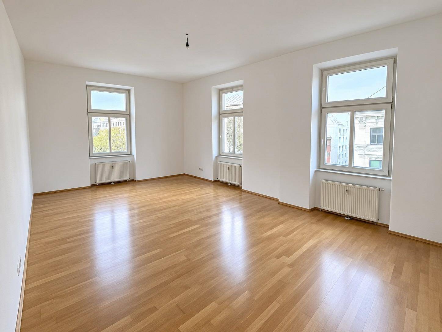 Helles und geräumiges Zimmer mit Parkettboden und drei großen Fenstern, ideal als Wohnbereich.
