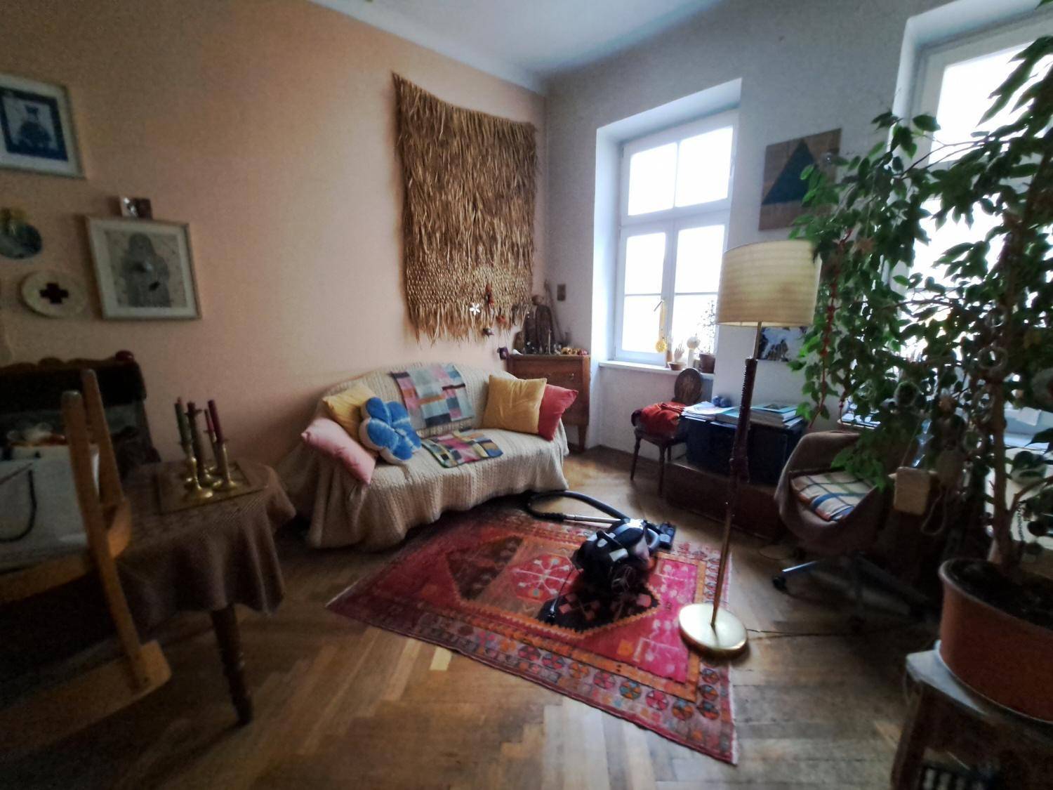 Gemütliches Wohnzimmer mit Holzboden, Sofa, Pflanzen und einem großen Fenster, das viel Licht hereinlässt.