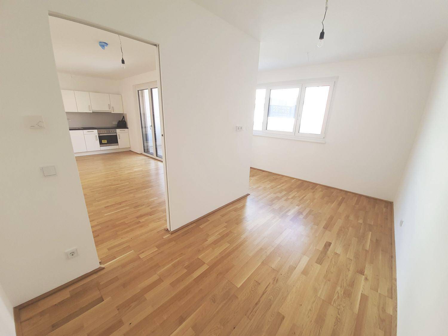 Geräumiger Wohnbereich mit Holzboden und Fenster, offen zur Küchenzeile und weiteren Räumen.