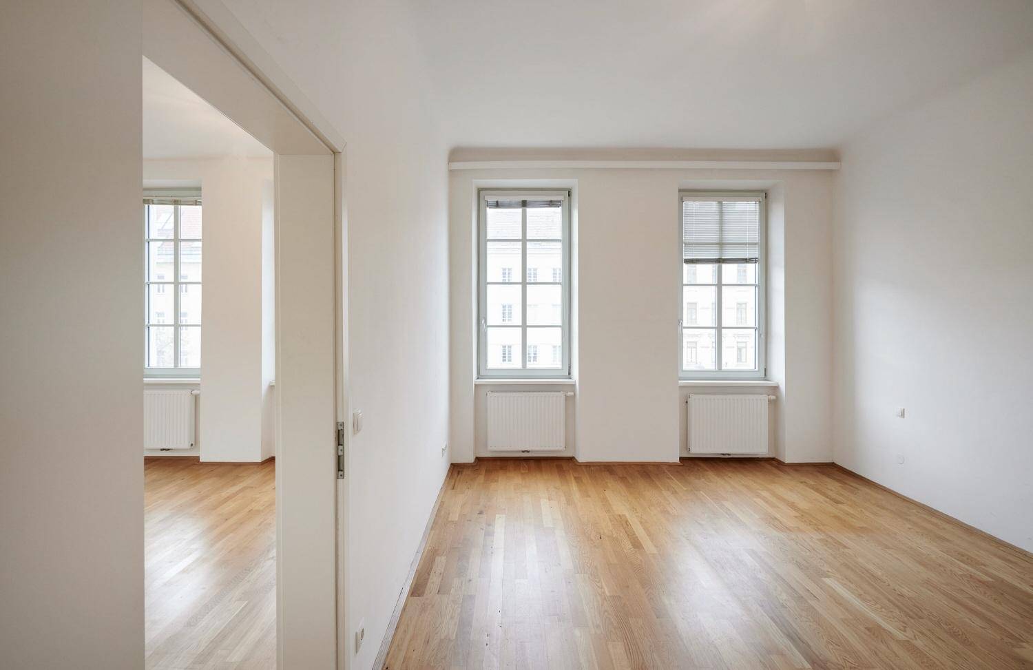 Geräumiges Zimmer mit Holzboden und zwei großen Fenstern, verbunden mit einem weiteren Raum.