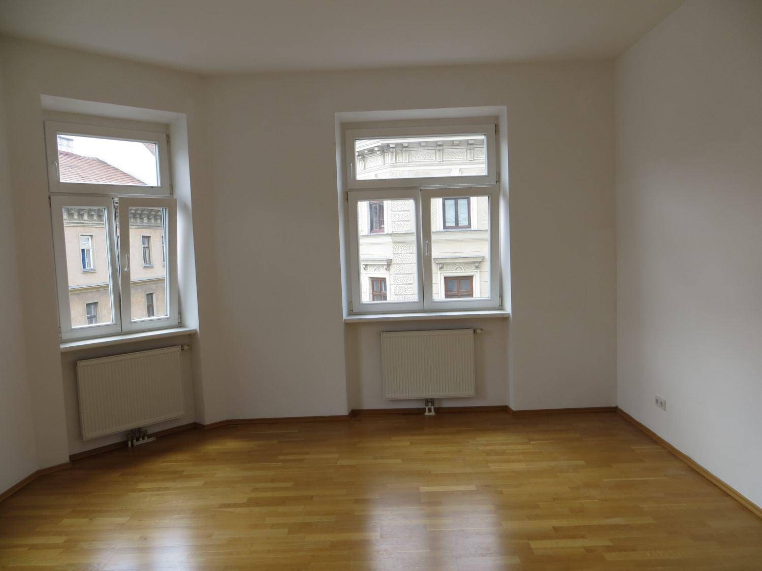 Lichtdurchflutetes Zimmer mit Parkettboden und zwei Fenstern, die auf die Straße blicken.
