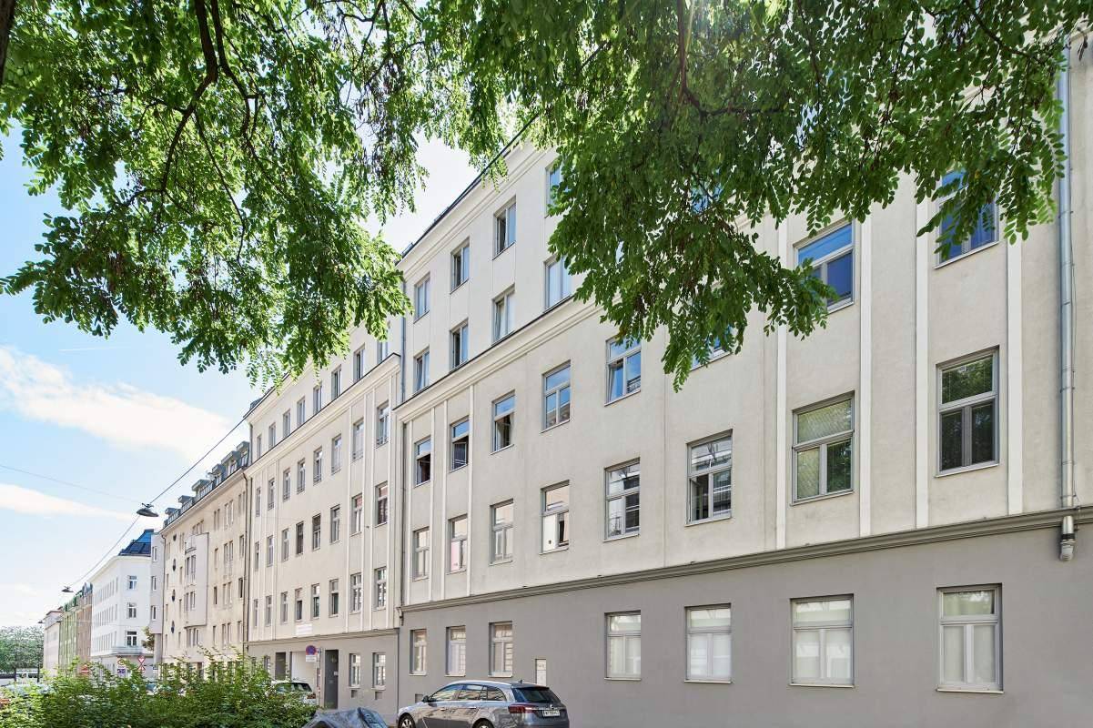 Seitenansicht eines Wohngebäudes mit vielen Fenstern und Bäumen im Vordergrund, bei Sonnenschein.