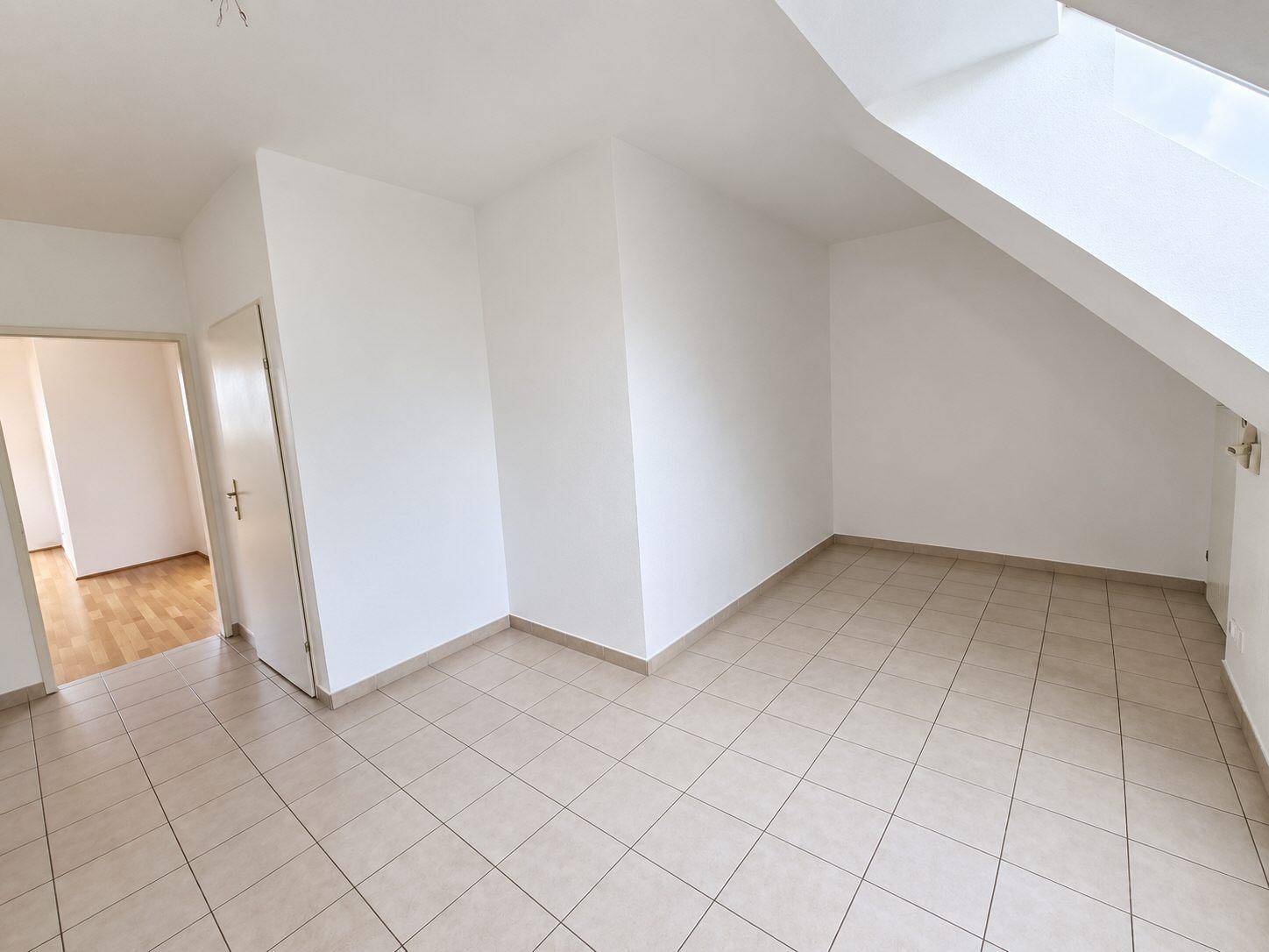 Heller Raum mit Fliesenboden und schrägen Wänden, der zu einem weiteren Zimmer führt.