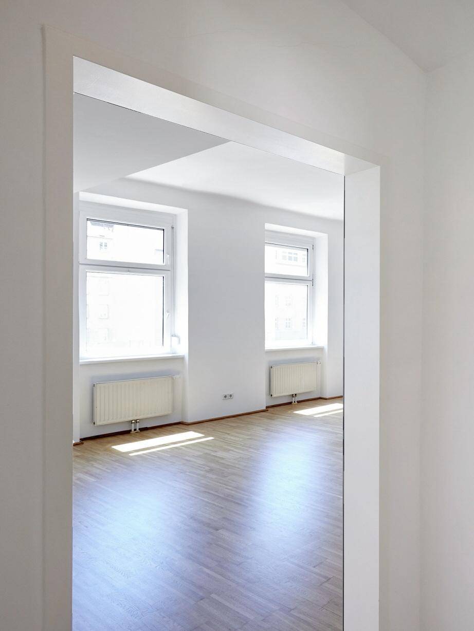 Blick durch einen Türrahmen in ein helles Zimmer mit Parkettboden und großen Fenstern.
