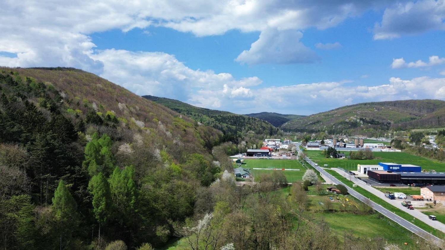 Luftaufnahme eines Tals mit einer Straße, Gebäuden und bewaldeten Hügeln in der Ferne.